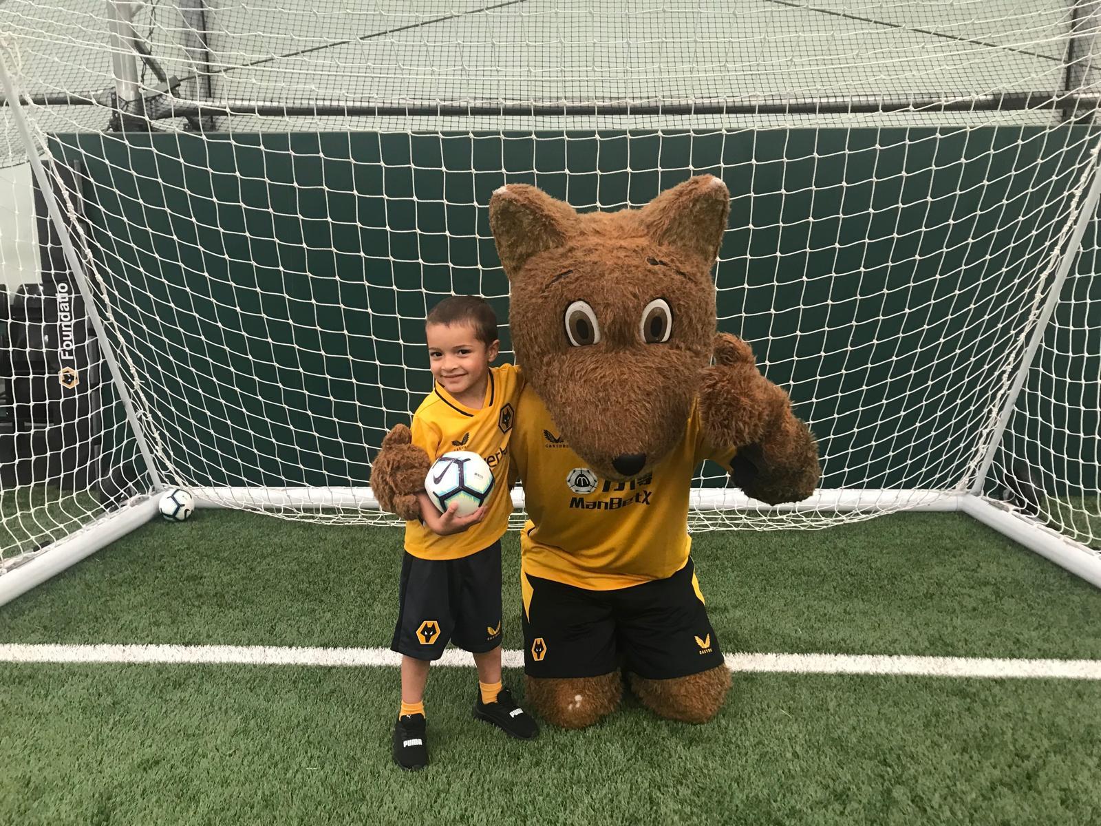 Xander in a gold home kit holding a ball in front of a goal beside the club mascot