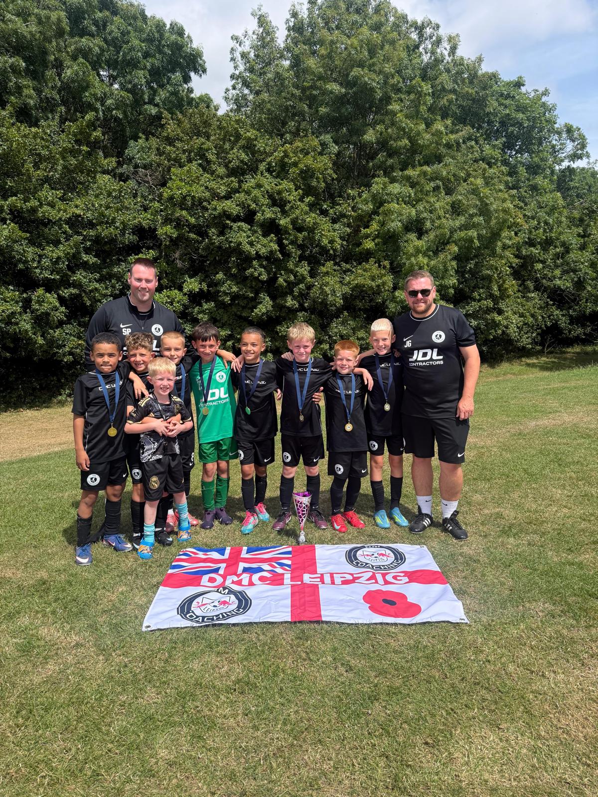 The DMC Leipzig squad and two coaches with medals behind their flag on a grass pitch