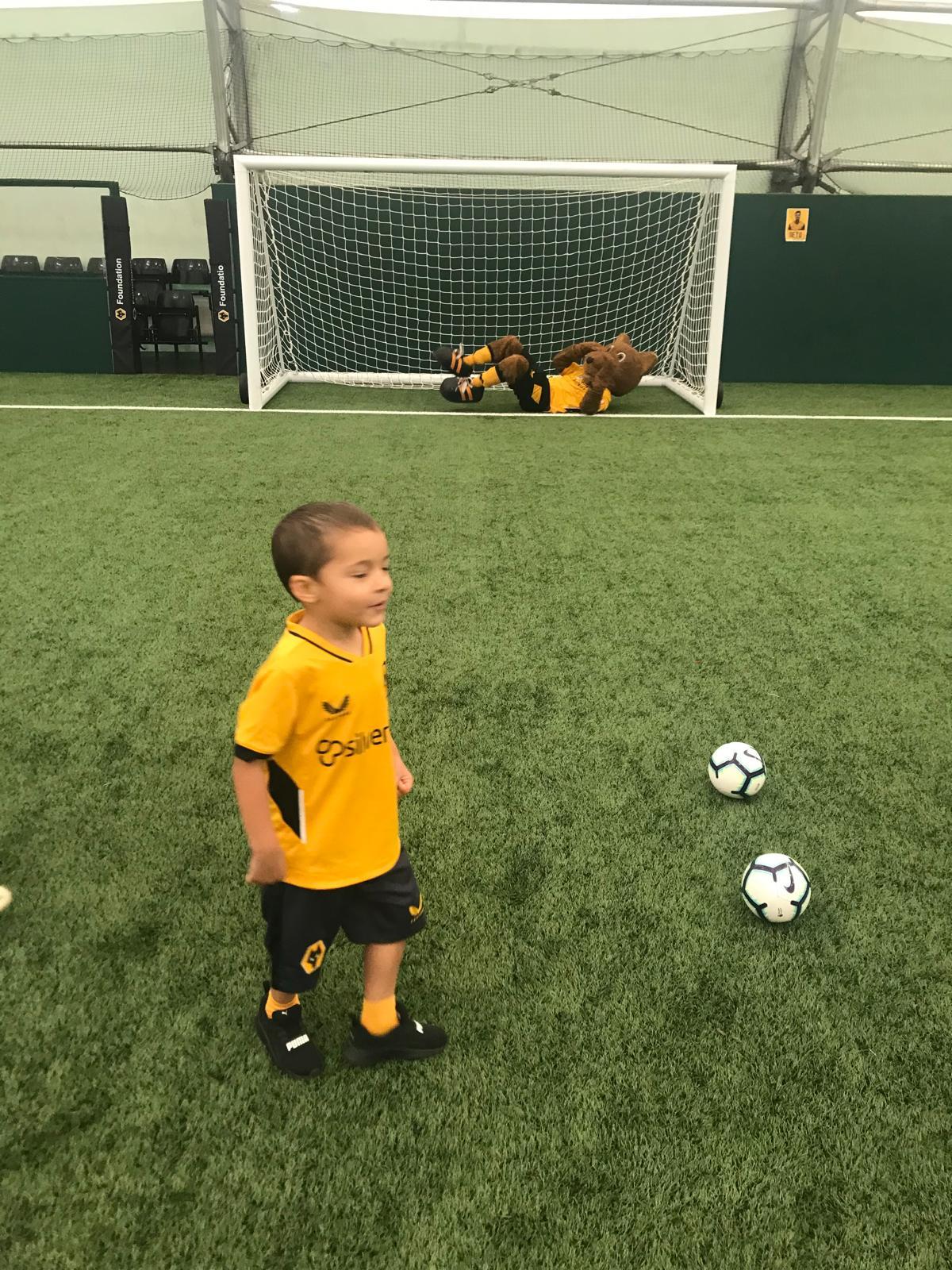 Xander in a gold and black kit on an indoor pitch with a club mascot in goal