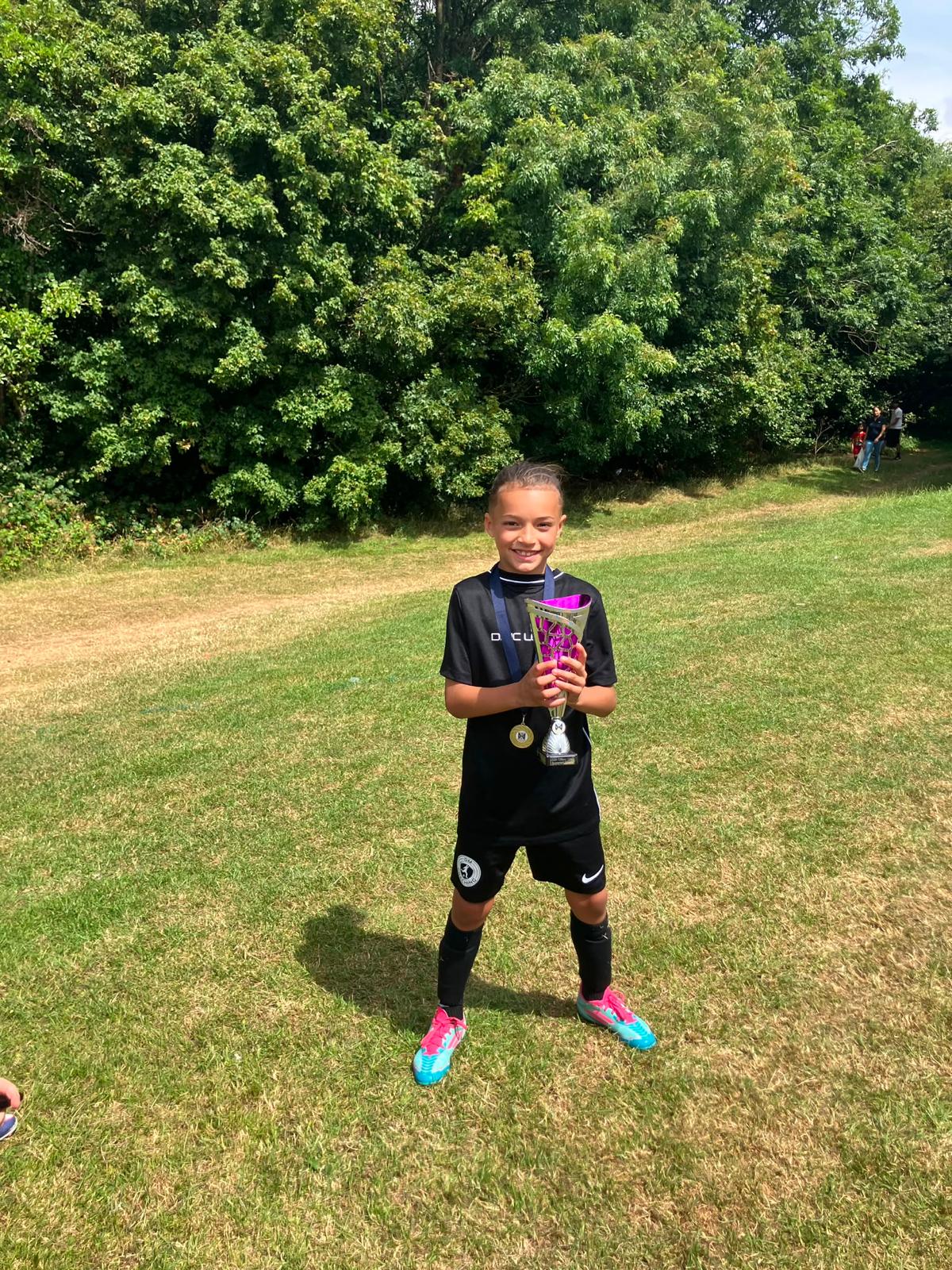 Xander in a black kit holding a trophy and medal on a sunny pitch